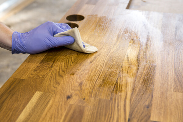 Hand wearing glove applying a clear topcoat to stained wood with a cloth.