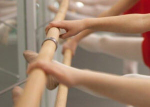 Two dancers using a smooth wooden ballet barre mounted to the wall in a studio setting.