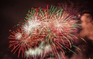 Bright aerial fireworks bursting above water from mortar tubes during a professional display.